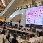 Busy shopping mall atrium with a large pink digital screen advertising a kids’ cosmetics experience school; attendees sit at tables for workshops.